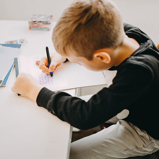 Jongen met een zwarte trui en grijze broek schrijft met een blauwe pen in een schrift.