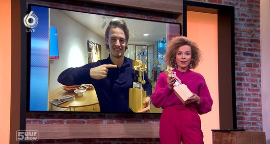 Man in marineblauw shirt wijst naar een houten flessenpuzzel op een groot scherm. Vrouw in roze outfit houdt een geschenkdoos vast.