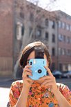 Een vrouw met een oranje gebloemd shirt houdt een lichtblauwe Instax Mini 11 camera vast.