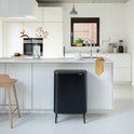 Poubelle Brabantia noire sur quatre pieds dans une cuisine moderne, à côté d'un tabouret de bar en bois.