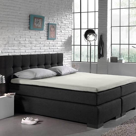 Donkergrijs bed met gestoffeerd hoofdbord, crème matras en twee kussens, in slaapkamer met witte muur.