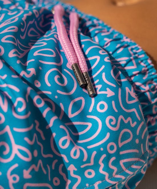 Detail van een turquoise zwembroek met een speels wit tekenpatroon en roze touwtjes met merkopdruk.