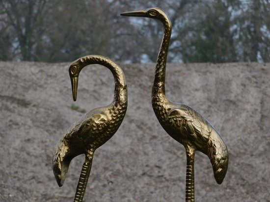 Deux statues de grues dorées, une avec le cou droit, l'autre avec le cou courbé, sur fond de sable et d'arbres.