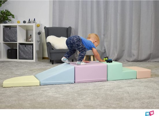 Un jeune enfant grimpe sur un ensemble de jeu souple aux couleurs pastel, comprenant une rampe jaune et des blocs bleu pastel, rose et menthe.