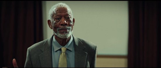 Morgan Freeman met grijs haar en baard, draagt een grijs pak, lichtblauw overhemd en gele stropdas.