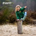 Kind in een groene pluizige jas en jeans, op een houten paal. Draagt groene laarzen.