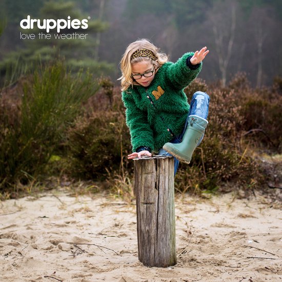 Kind in een groene pluizige jas en jeans, op een houten paal. Draagt groene laarzen.