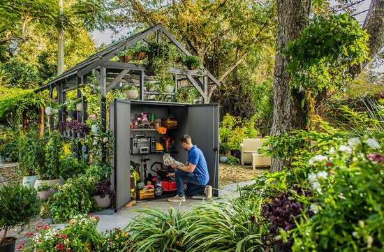 Homme ouvrant un abri de jardin gris dans un jardin luxuriant, avec des outils et des plantes à l'intérieur.