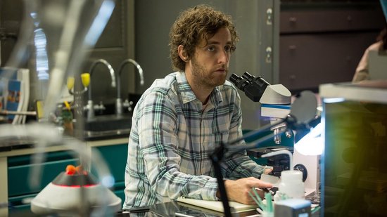 Een man met krullend haar kijkt door een microscoop in een laboratorium.