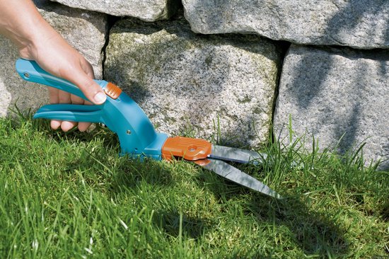 Hand gebruikt blauwe Gardena graschaar om gras te knippen langs stenen muur.