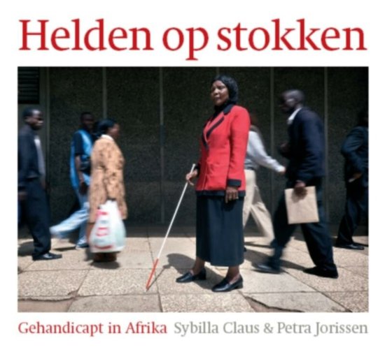Vrouw in rode jas en zwarte rok, met witte stok in de hand, staand op trottoir.