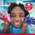 Een jong kind met verf op de handen, lachend naar de camera. Het kind heeft rood, geel en blauwe verf op zijn handen.