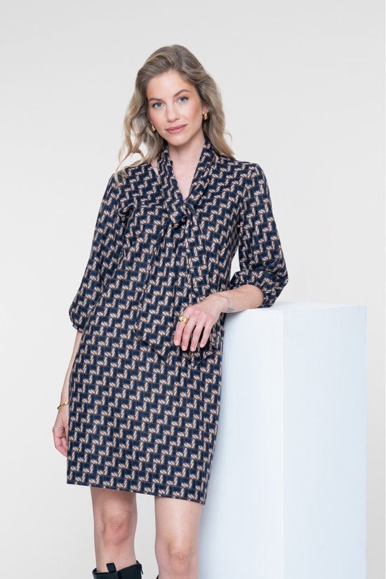 Vrouw met blonde haren, draagt een navy jurk met geometrische print en strikdetail, met gouden accessoires.
