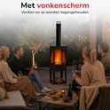 Zwarte buitenhaard met schoorsteen en vonkenscherm, met brandend vuur. Groep mensen geniet op terras.