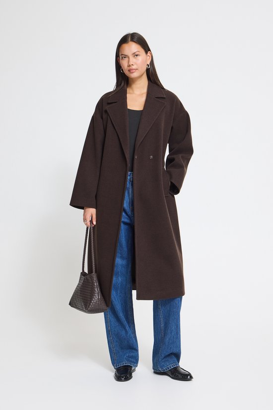 Model draagt een donkerbruine jas, blauwe jeans en draagt een gevlochten tas.