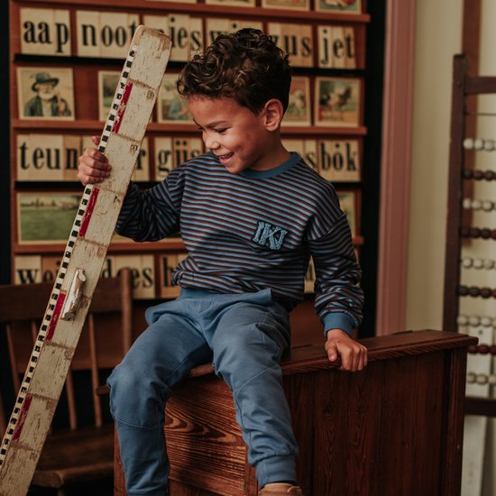 Jongen met lachend gezicht, gekleed in een blauw-bruin gestreepte trui met het embleem 'IK' en een blauwe joggingbroek.