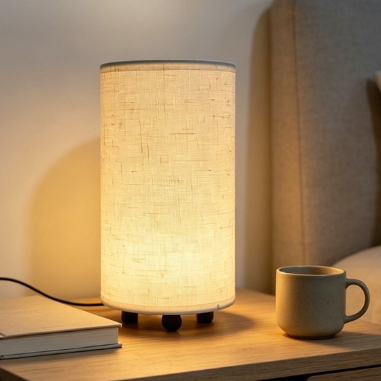 Cilindervormige beige linnen tafellamp op een nachtkastje, naast een boek en een mok. Warm en sfeervol licht.
