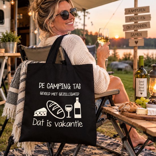Sac noir avec le texte 'DE CAMPING TAS GEVULD MET GEZELLIGHEID Dat is vakantie' et des images de fromage, pain et vin. Une femme est assise avec le sac sur ses genoux.