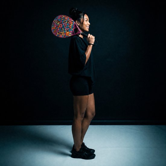 Vrouw met donker haar in een knot, lachend, die een padelracket met een kleurrijk cartoonpatroon op haar schouder draagt. Ze draagt een zwarte T-shirt en korte broek.