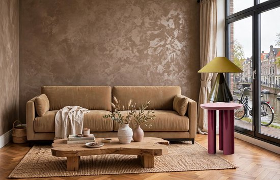 Een taupe bank met een verweerde textuur muur erachter. Een houten salontafel met een vloerkleed, lamp en bijzettafel staan ervoor.