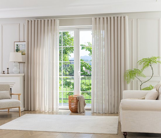 Zandkleurige vitragegordijnen hangen voor een raam met uitzicht op groen gebladerte en een balkon. Een fauteuil en bank zijn in de kamer.