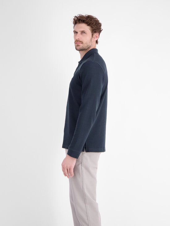 Zijaanzicht van een model in een marineblauw poloshirt met lange mouwen en een grijsbeige broek.
