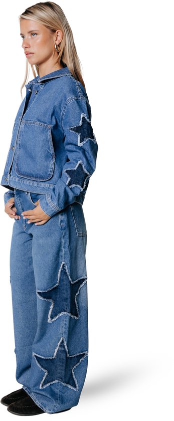 Blauw denim jack met sterapplicaties op de mouwen, gedragen door een model met een spijkerbroek.
