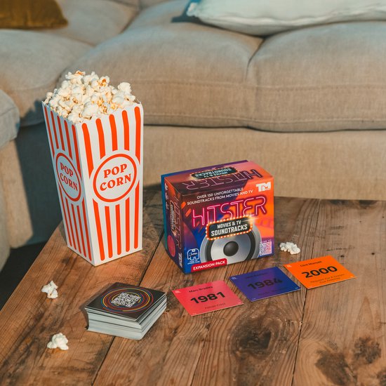 Hitster 'Movies & TV Soundtracks' uitbreidingsset op een houten tafel met een bak popcorn en speelkaarten met jaartallen.