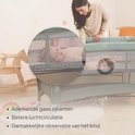 Moeder kijkt naar baby in een groen Lionelo reisbedje met ademende gaaszijkanten voor betere luchtcirculatie.