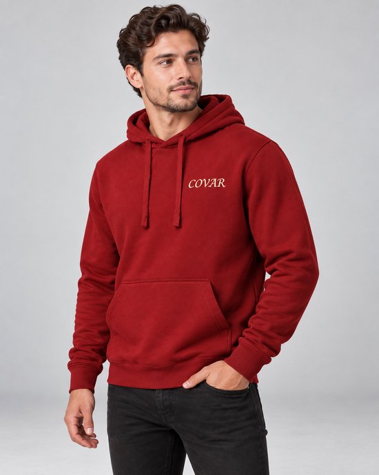 Man met donkere jeans, witte sneakers en rode COVAR hoodie met capuchon, staand.
