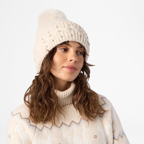 Model draagt een beige gebreide muts met strass-steentjes en een pluizige pompon.