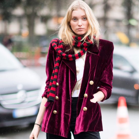 Close-up van een model in een wijnrode fluwelen blazer met gouden knopen en een rood-zwarte gestreepte sjaal.