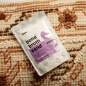 Pakje 'bone broth eend' met een paarse eend erop, op een textiel met bruine en beige patronen.