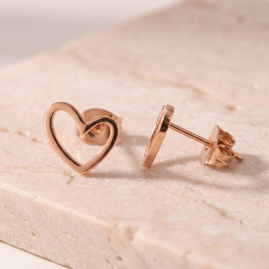 Twee rosegoudkleurige hartjesoorbellen met opengewerkte vorm, liggend op een lichtroze steen.