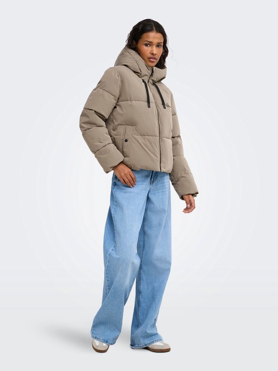 Model draagt een beige, gewatteerde pufferjas met capuchon, zwarte ritssluiting en lichtblauwe wijde jeans.