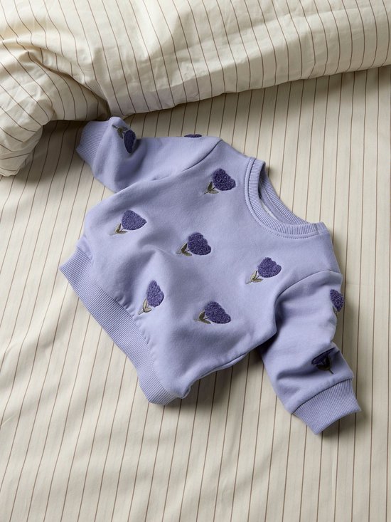 Lichtpaarse sweater met hartvormige bloemmotieven, liggend op gestreept beddengoed.