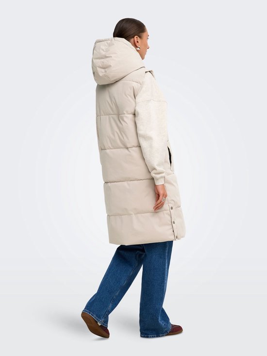 Achterzijaanzicht van een model met een lange beige bodywarmer met capuchon en spijkerbroek.