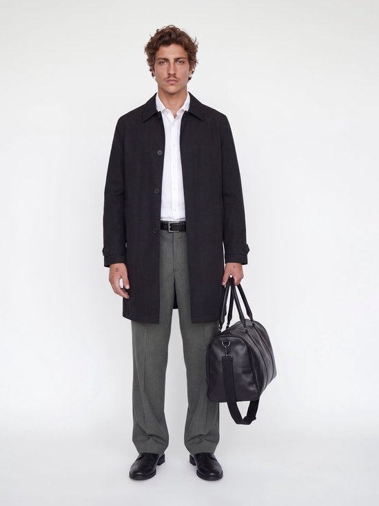 Man met witte hemd, grijze wijde broek, zwarte riem, zwart jasje en zwarte weekendtas.