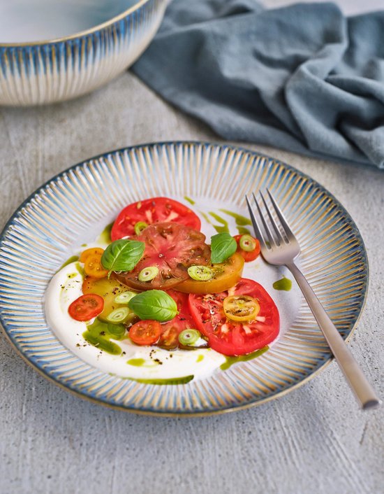 Diep bord met blauw-bruin gestreept patroon, gevuld met een tomatensalade met basilicum en een vork op de rand.