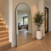 Grote boogspiegel met slank zwart frame, staand in een modern interieur naast een trap en een plant in een pot.