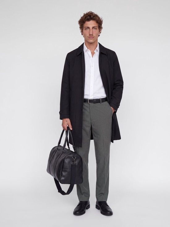 Man met bruin krullend haar en snor, gekleed in een wit overhemd, grijze wijde broek, zwarte riem, zwarte jas en zwarte schoenen. Hij draagt een zwarte weekendtas.