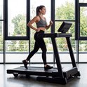 Vrouw met een VirtuFit hartslagmeter om haar borst, gekleed in sportkleding, traint op een loopband in een fitnessruimte.