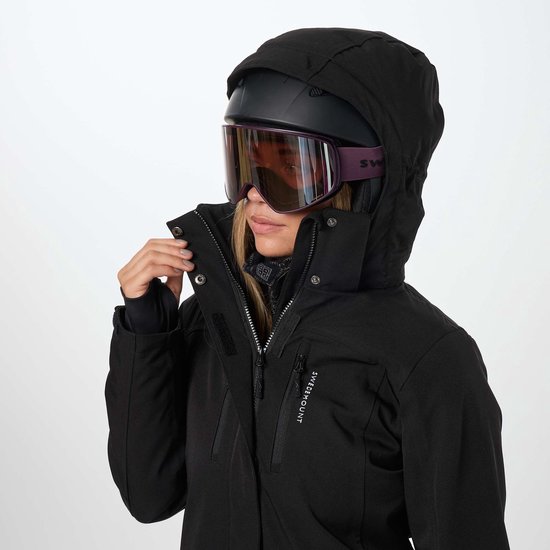 Close-up van een vrouw die haar zwarte ski-jas sluit, met paarse skibril en helm.
