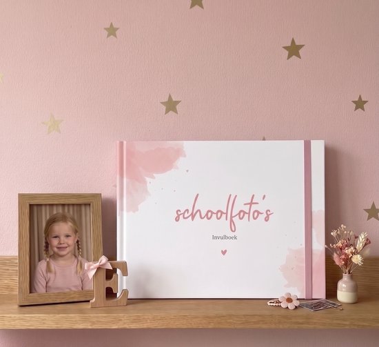 Wit invulboek voor schoolfoto's op een houten plank naast een fotolijstje van een meisje en droogbloemen.
