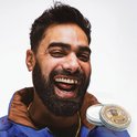 Lachende man met korte baard en blauwe jas, naast een blikje Reuzel baardbalsem.