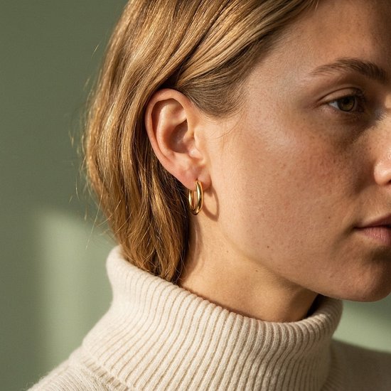 Model draagt een kleine, glanzende gouden oorring van de Lundholm Laura-serie bij een beige coltrui.