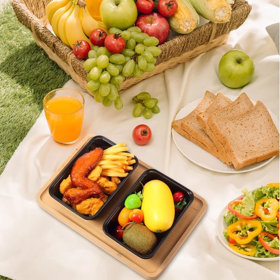 Picknick met zwarte voedselcontainers, fruitschaal, brood, salade en sap, op een kleed in het gras.