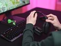 Iemand houdt een zwarte bedrade gamingheadset vast in een gamingopstelling met een toetsenbord en monitor.