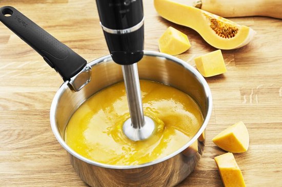 Zwarte Tefal staafmixer in een pan met oranje soep, naast stukken pompoen op een houten ondergrond.