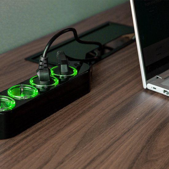 Zwarte stroomstrip met verlichte groene stopcontacten, aangesloten op een laptop, op een houten bureau.